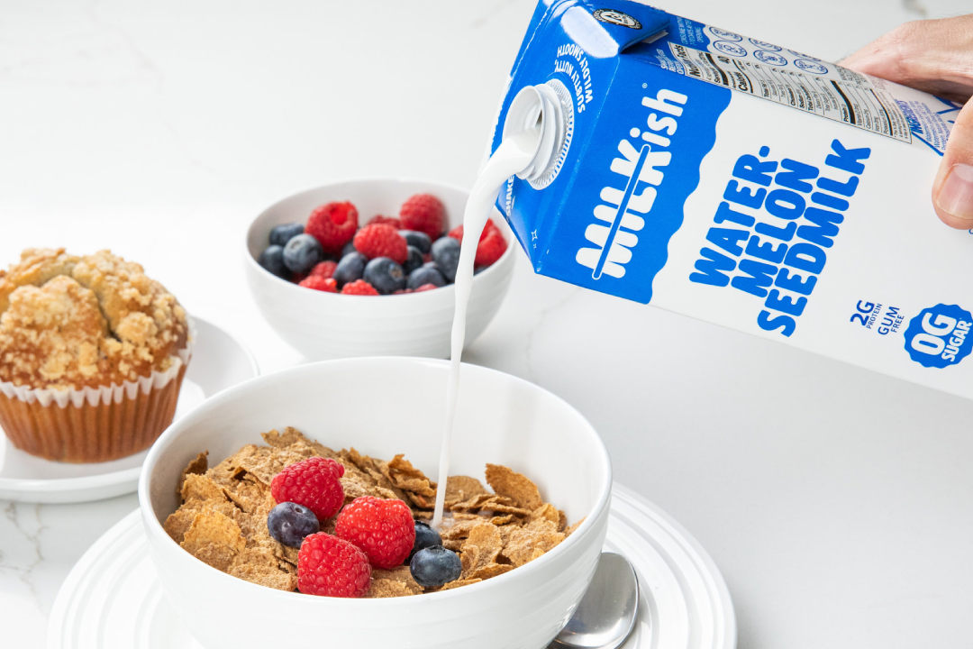 Person pouring milk from a carton labeled 'Watermelon Seedmilk' into a bowl of cereal with berries on a white surface.