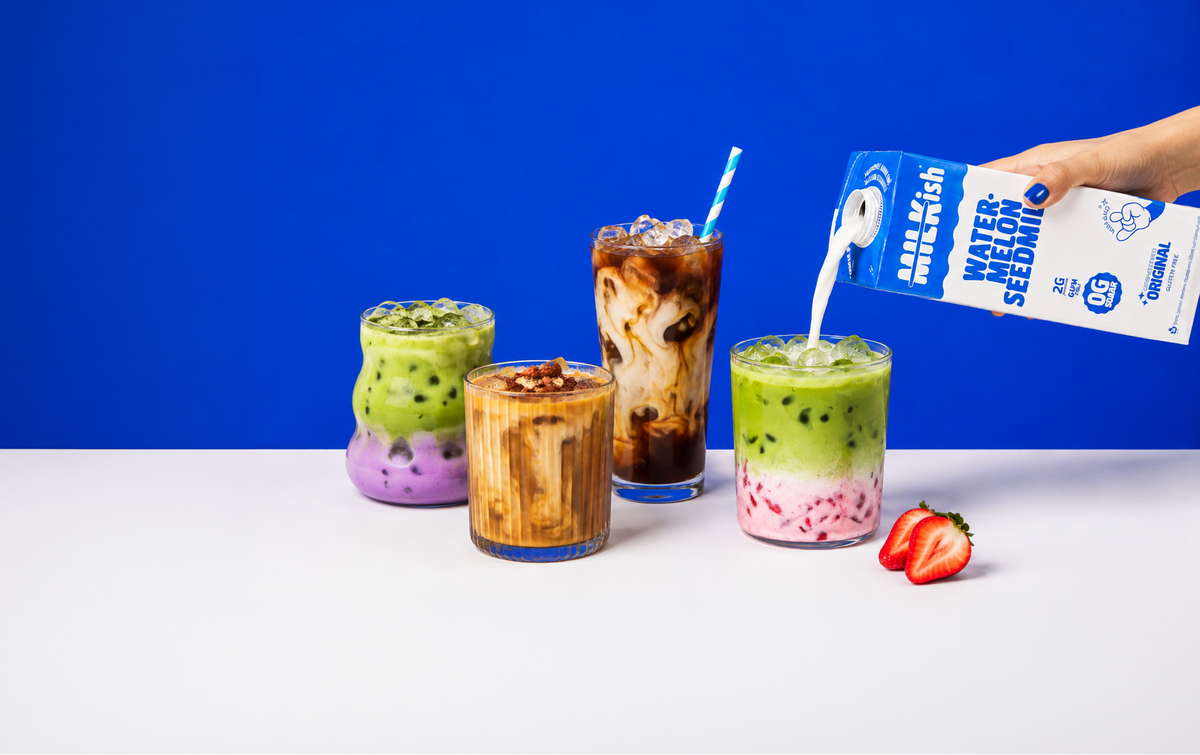 Four colorful layered drinks on a white surface against a solid blue background, with a hand pouring watermelon seed milk from a blue carton into one of the glasses. Strawberries rest beside the drinks.
